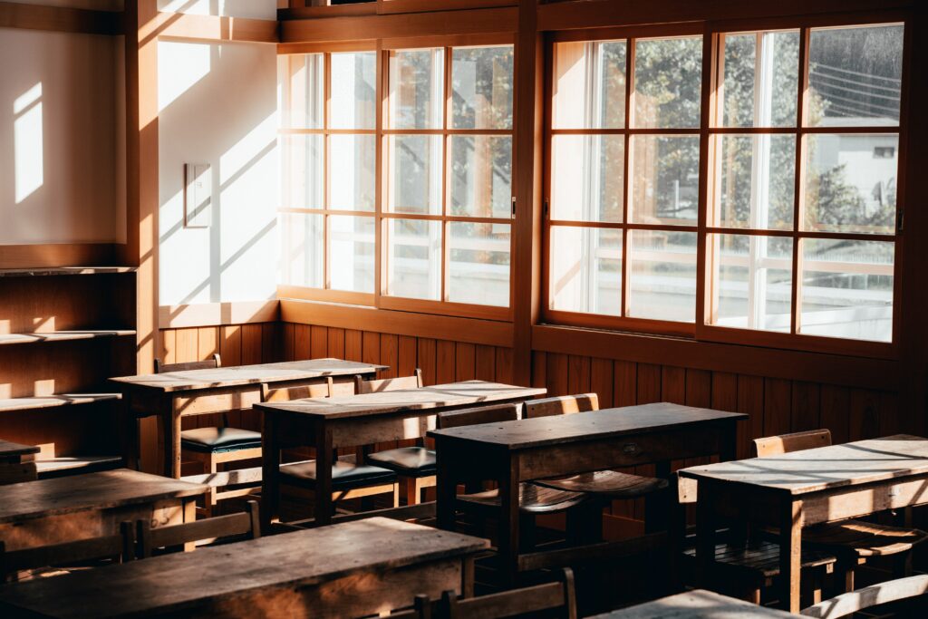 Empty Japanese classroom with four rows of two-person wood desks surrounded by windows with sunlight shining in and wooden walls.